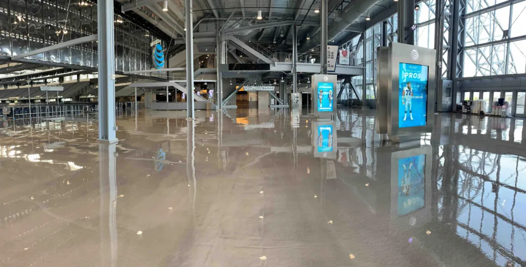 A spacious interior with resinous floors reflects overhead lighting and digital advertisements. Large windows on the right allow natural light into the area.