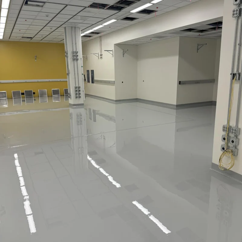 Large, empty room with a shiny, light gray epoxy floor, yellow and white walls, ceiling panels, and visible electrical conduits.