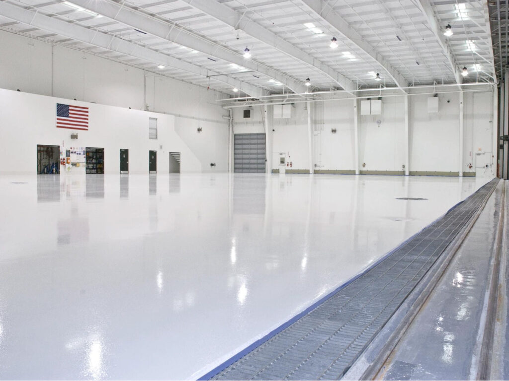 Large, empty, white indoor warehouse space with a polished floor, high ceiling, an American flag on the wall, and several doors along the back.