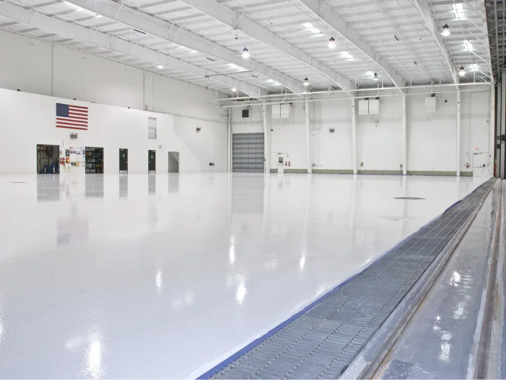 Large, empty, white indoor warehouse space with a polished floor, high ceiling, an American flag on the wall, and several doors along the back.