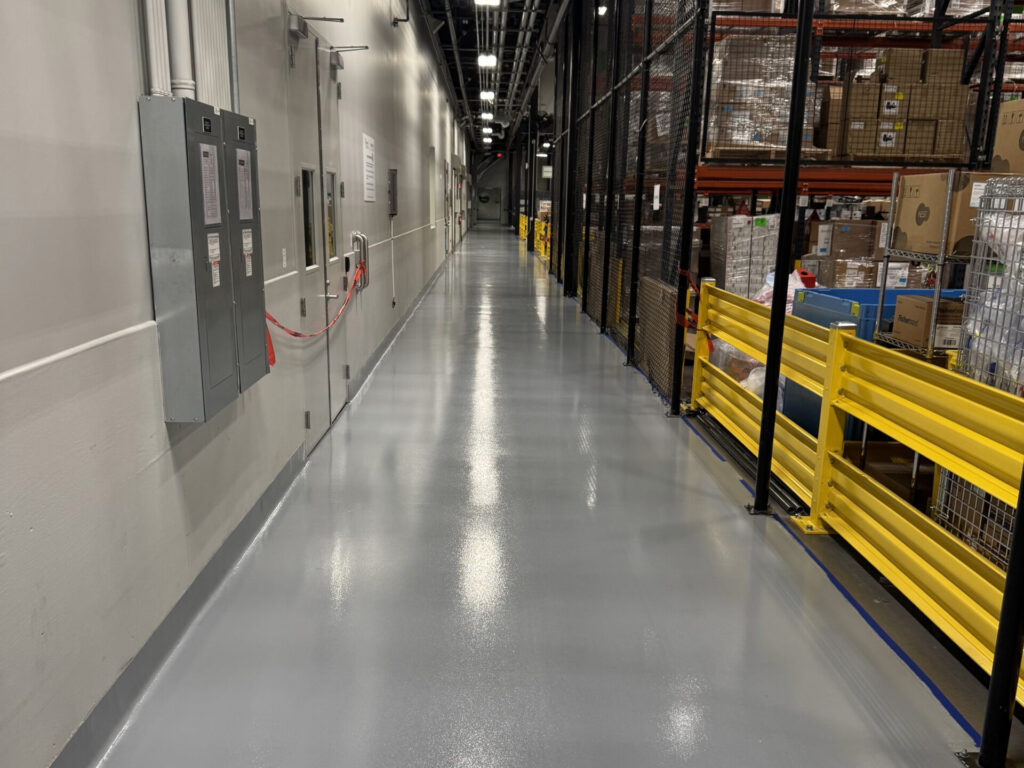 A clean, shiny hallway in an industrial facility with electrical panels on the left and storage racks with boxes on the right, separated by a yellow barrier.