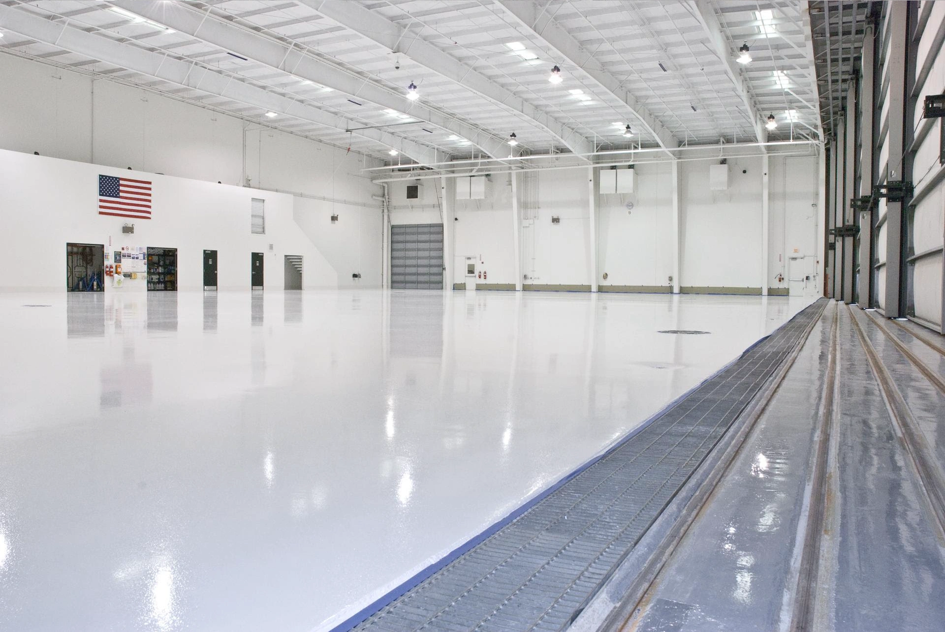 A large, empty hangar with a smooth, reflective floor. The interior is illuminated by overhead lights. An American flag hangs on the left wall.