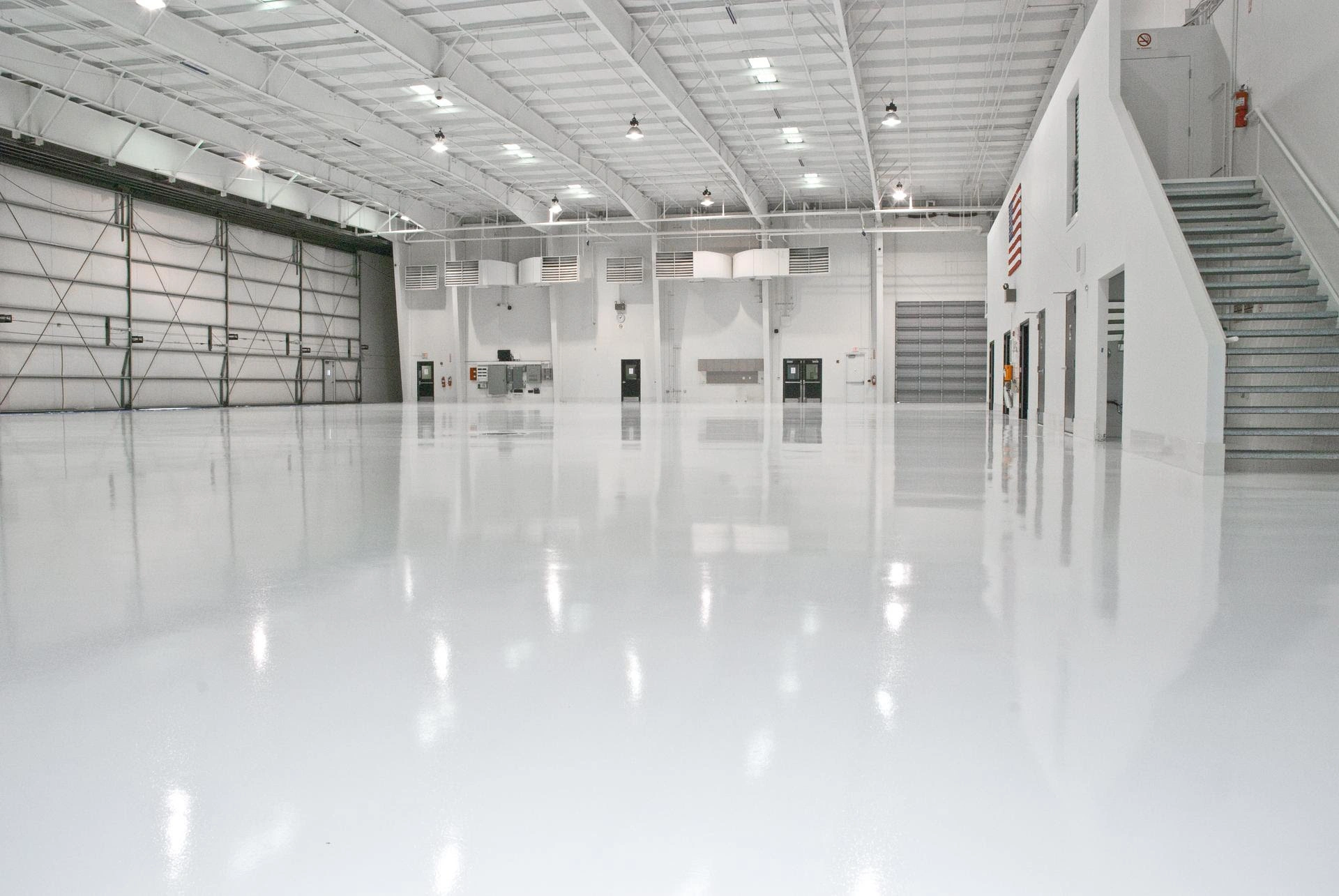 Spacious, empty airplane hangar with a glossy white floor, high ceiling, large overhead doors, and a staircase on the right.