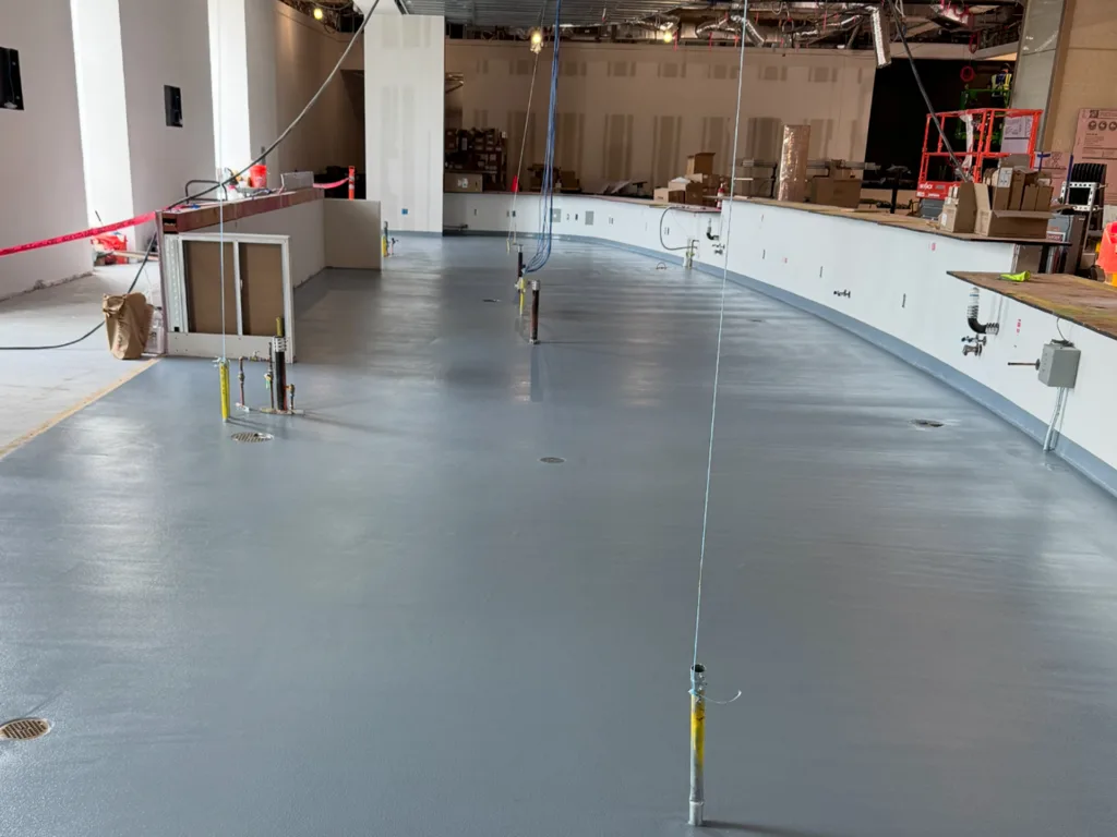 A large indoor space under construction with a smooth gray floor, wiring, exposed utilities, and boxes and equipment in the background.