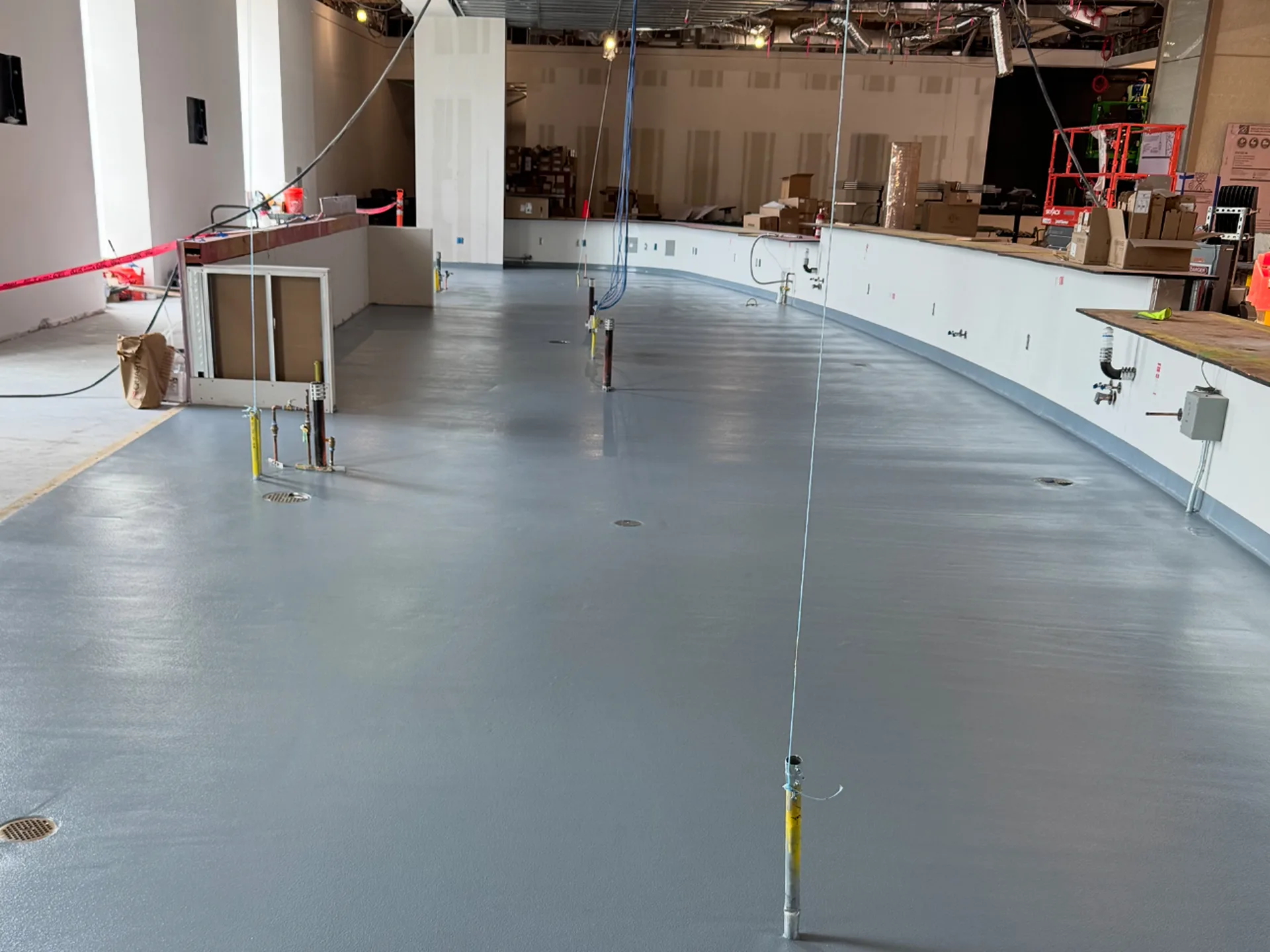 A large indoor space under construction with a smooth gray floor, wiring, exposed utilities, and boxes and equipment in the background.