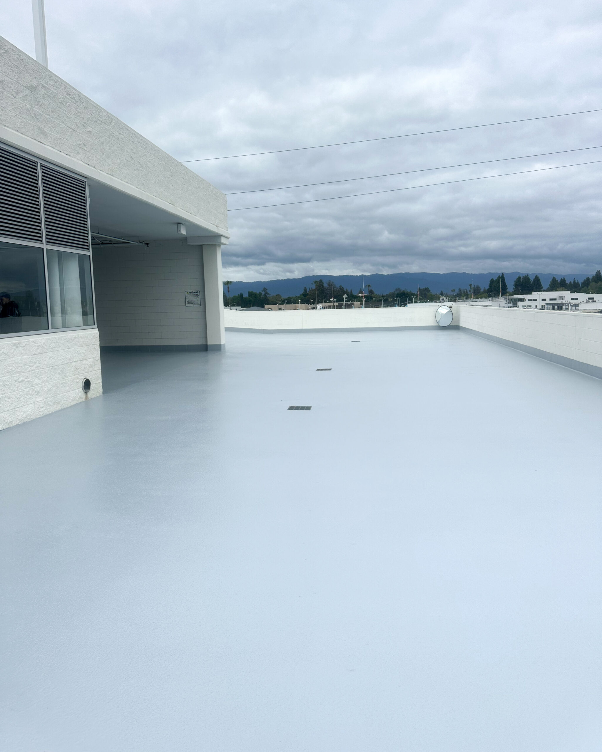 Flat, empty rooftop with a smooth, light grey surface, surrounded by low white walls, under an overcast sky with scattered clouds. Mountains and trees visible in the distance.