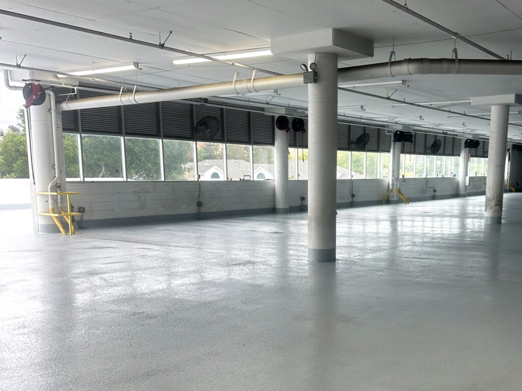 An empty, clean parking garage level with concrete floors, large windows along one wall, pillars, and overhead pipes visible.
