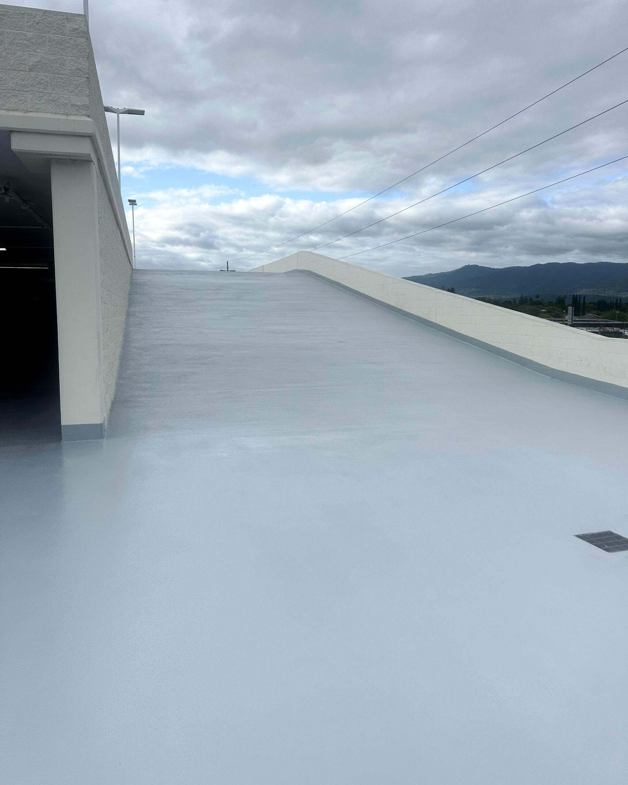 A clean, empty parking garage ramp with a smooth light gray surface, leading up to an overcast sky. Power lines and distant hills are visible in the background.