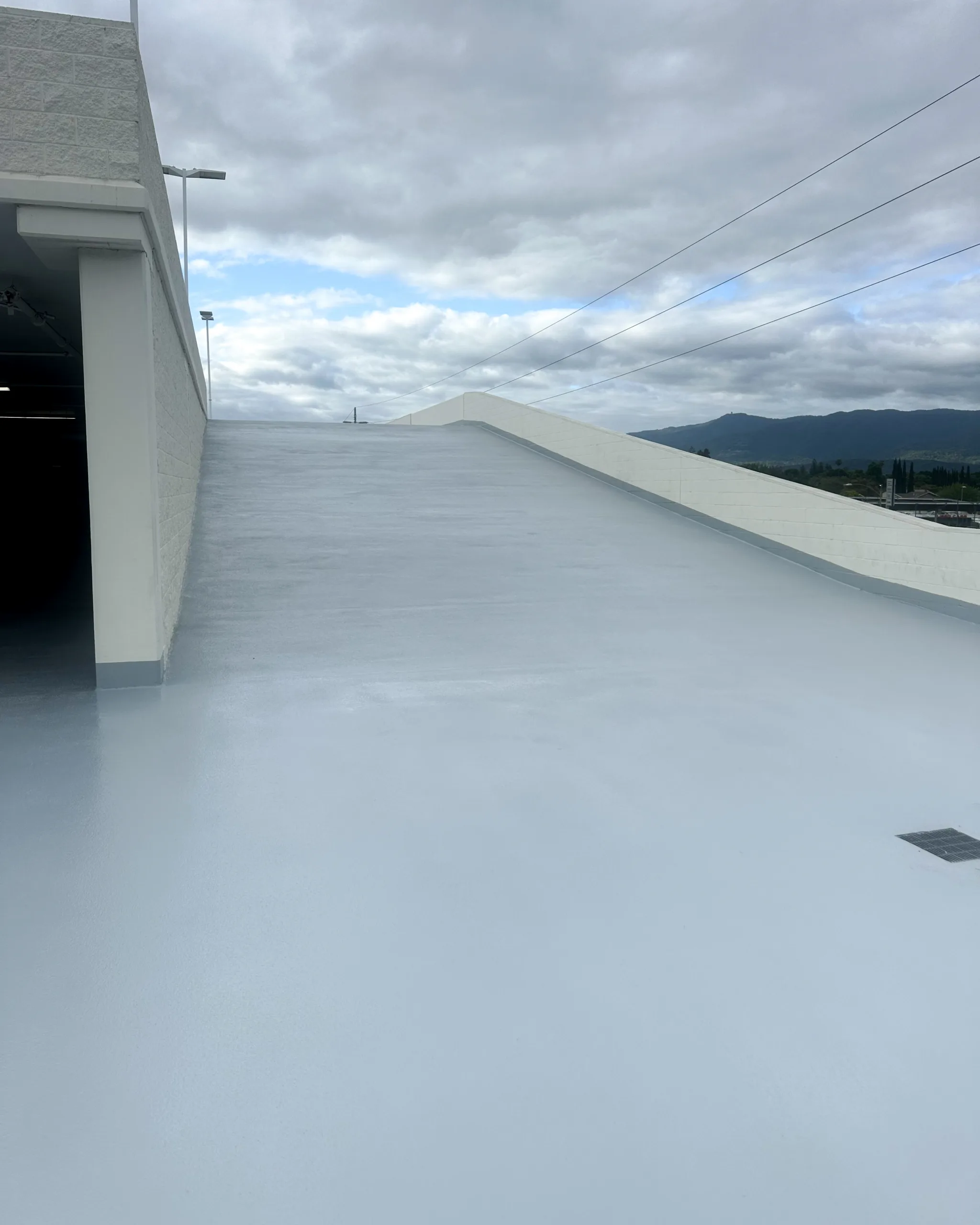 A clean, empty parking garage ramp with a smooth light gray surface, leading up to an overcast sky. Power lines and distant hills are visible in the background.