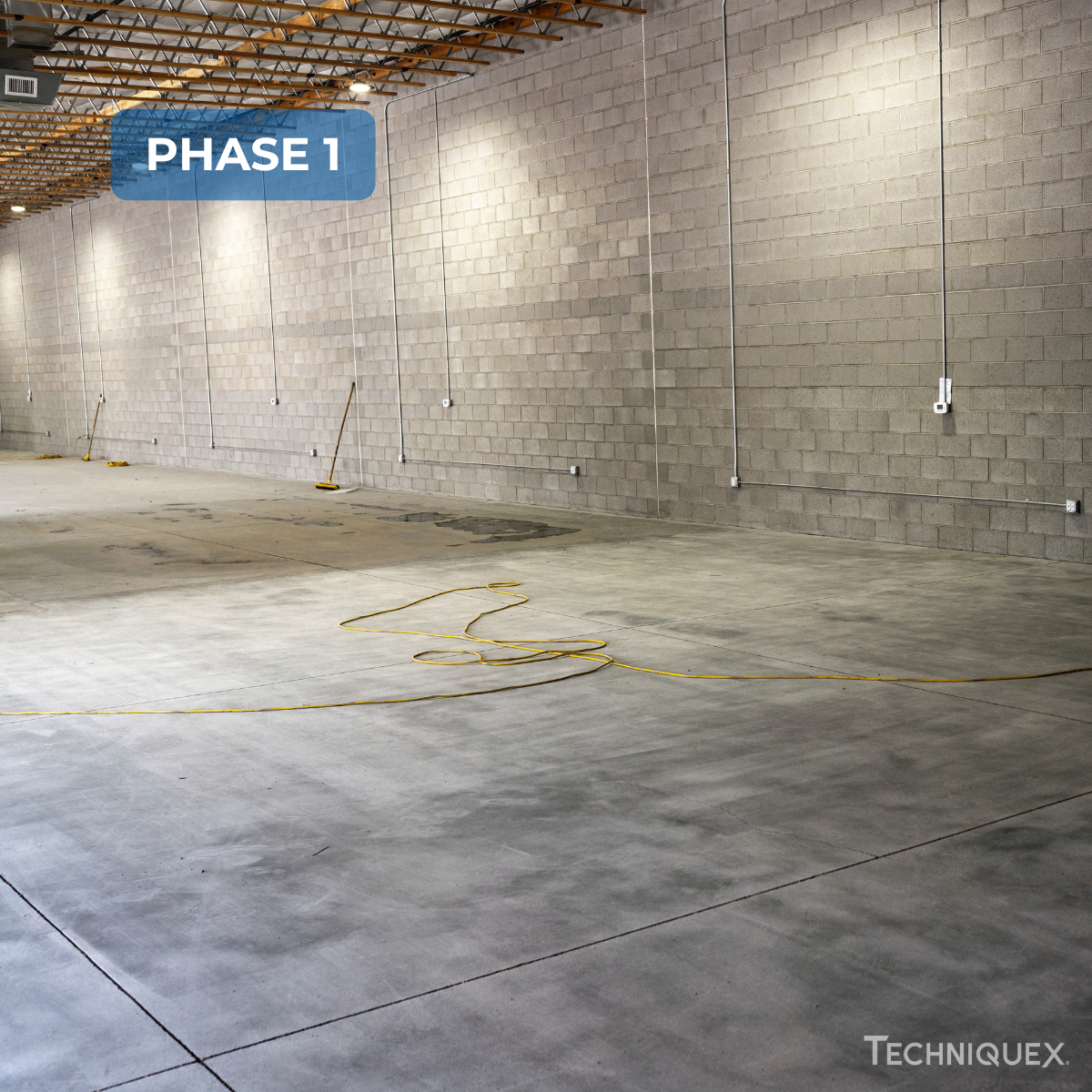 Large empty warehouse with concrete floors, cinder block walls, several brooms against the wall, and a yellow extension cord on the floor. Text reads "PHASE 1.