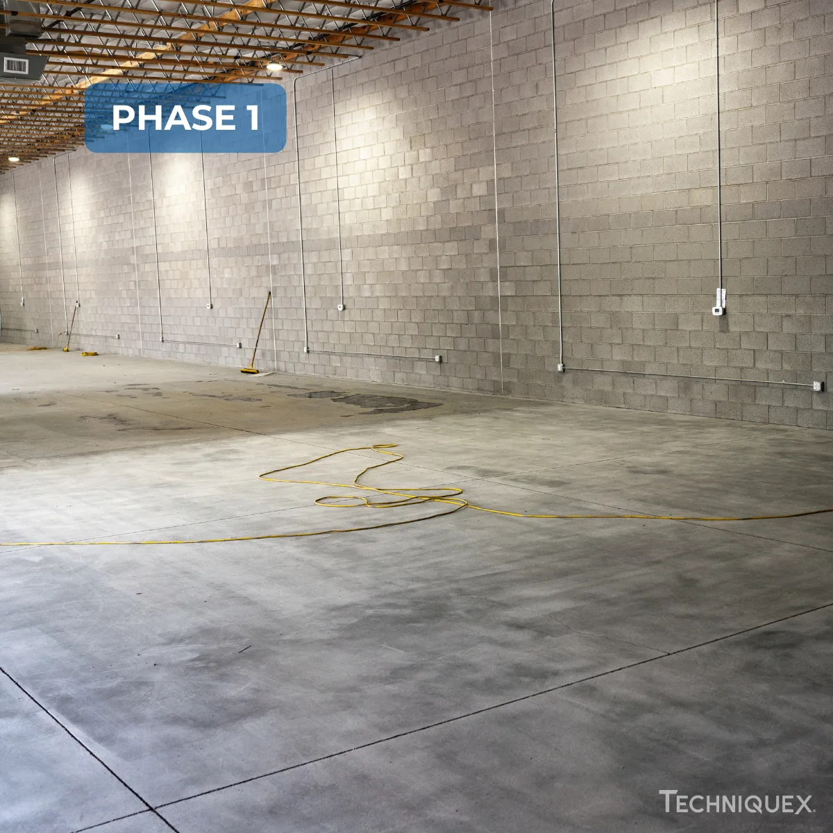 Large empty warehouse with concrete floors, cinder block walls, several brooms against the wall, and a yellow extension cord on the floor. Text reads "PHASE 1.