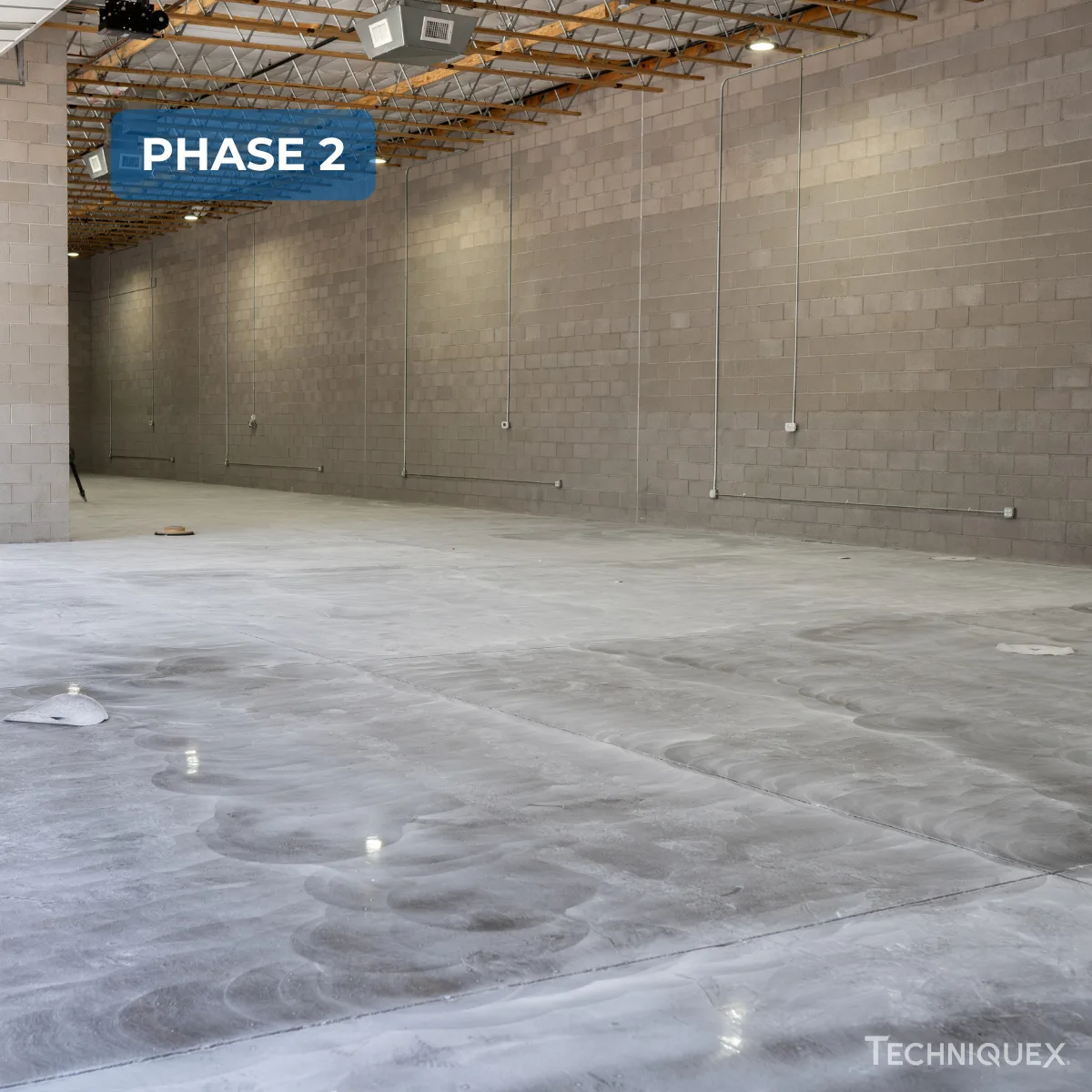 Large empty warehouse space with unfinished concrete floor, gray cinderblock walls, and exposed ceiling beams; text label reads "PHASE 2" in upper left.