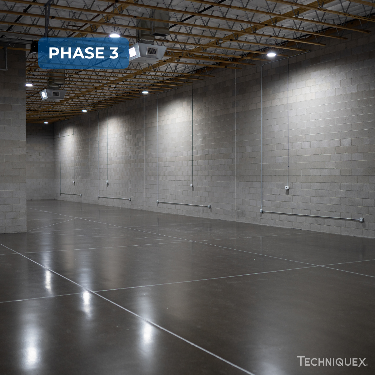 An empty industrial warehouse with concrete floors and cinder block walls, labeled "Phase 3," with overhead lighting and visible ceiling framework.