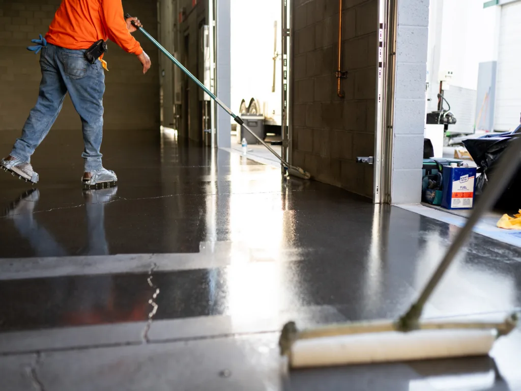 Person wearing an orange shirt applies a coating to a concrete floor with a roller in an industrial or commercial garage space.