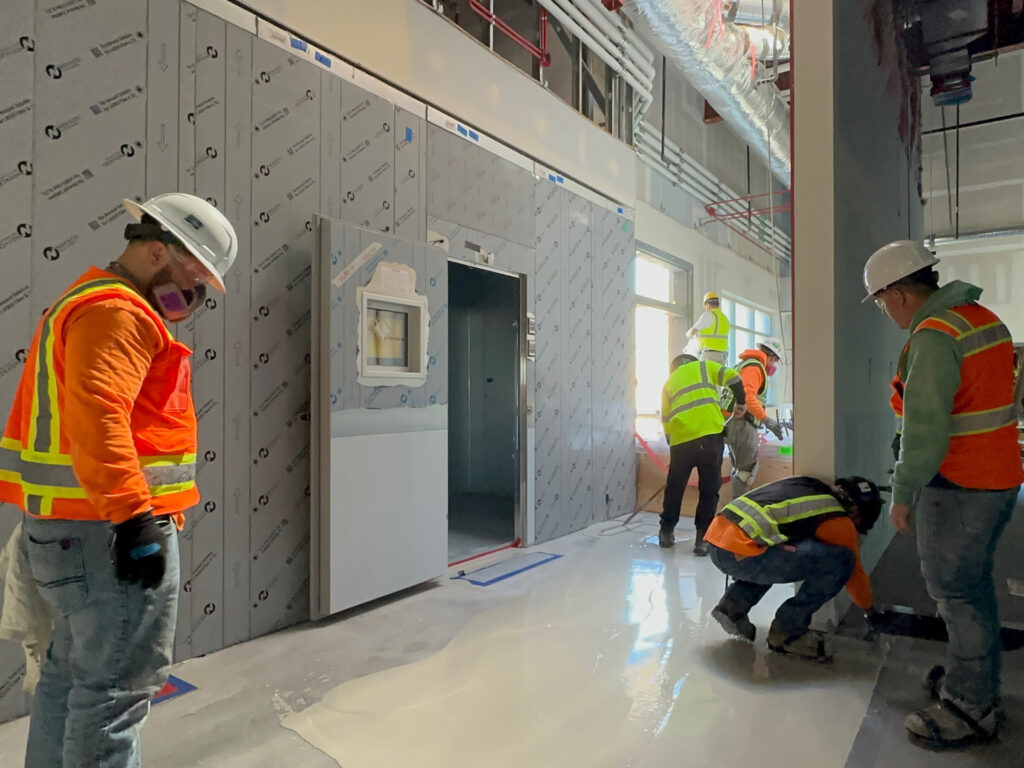 Construction workers in safety gear install flooring and inspect work inside a building under construction, with unfinished walls and visible pipes overhead.