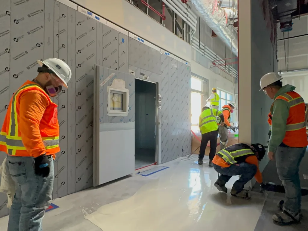 Construction workers in safety gear install flooring and inspect work inside a building under construction, with unfinished walls and visible pipes overhead.