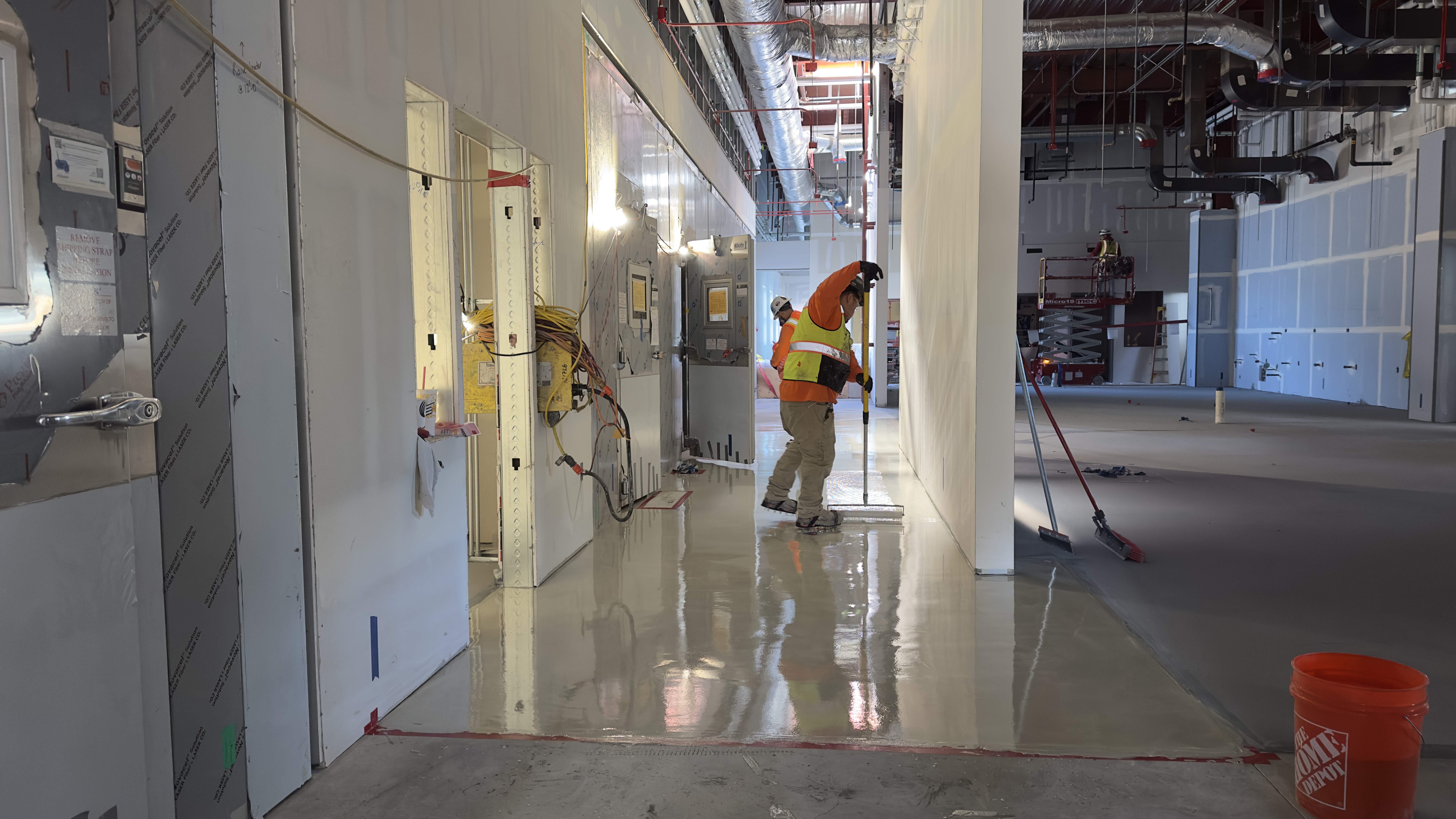 A construction worker in a safety vest and helmet spreads liquid material on the floor of an unfinished building interior.
