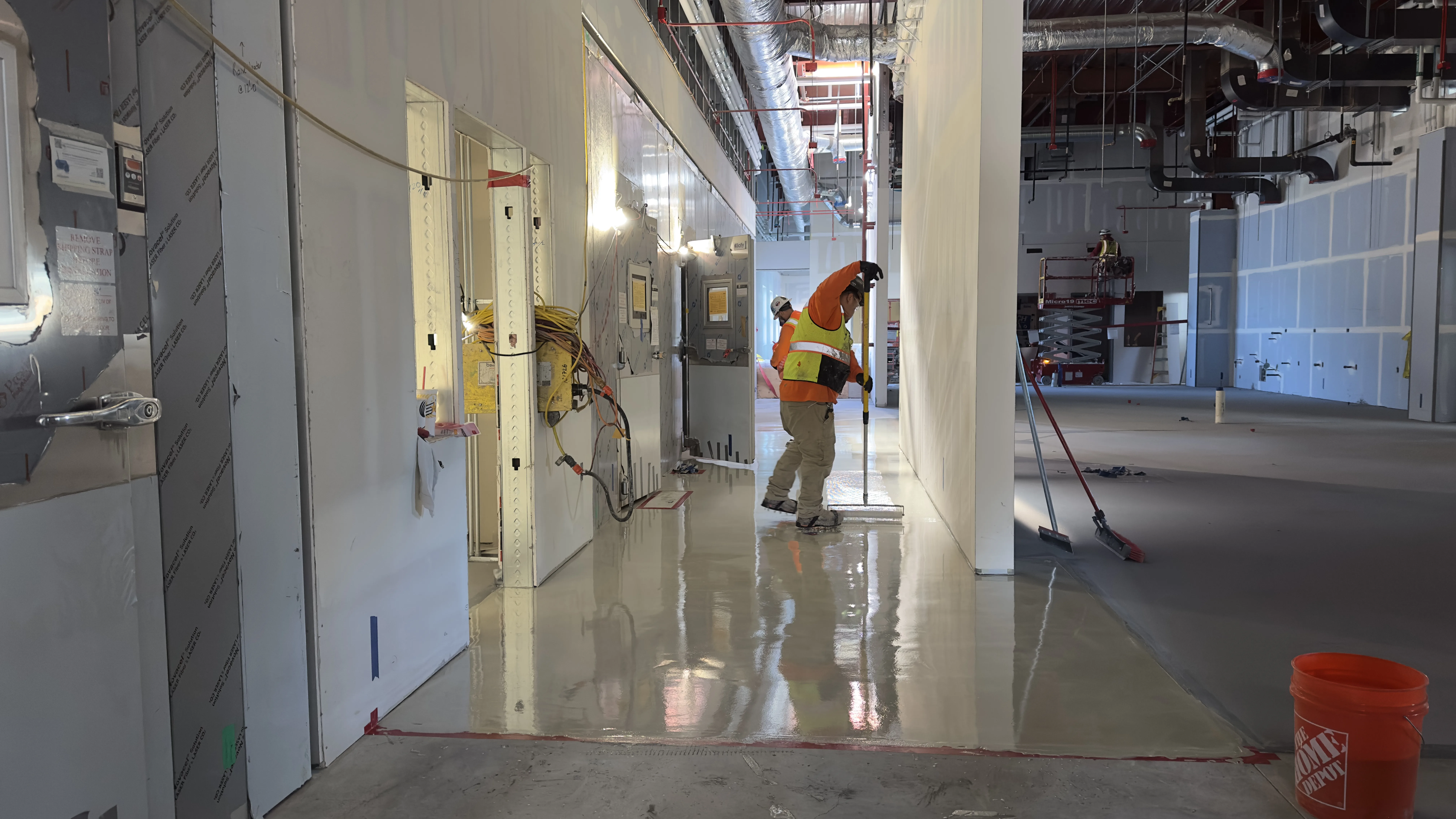 A construction worker in a safety vest and helmet spreads liquid material on the floor of an unfinished building interior.