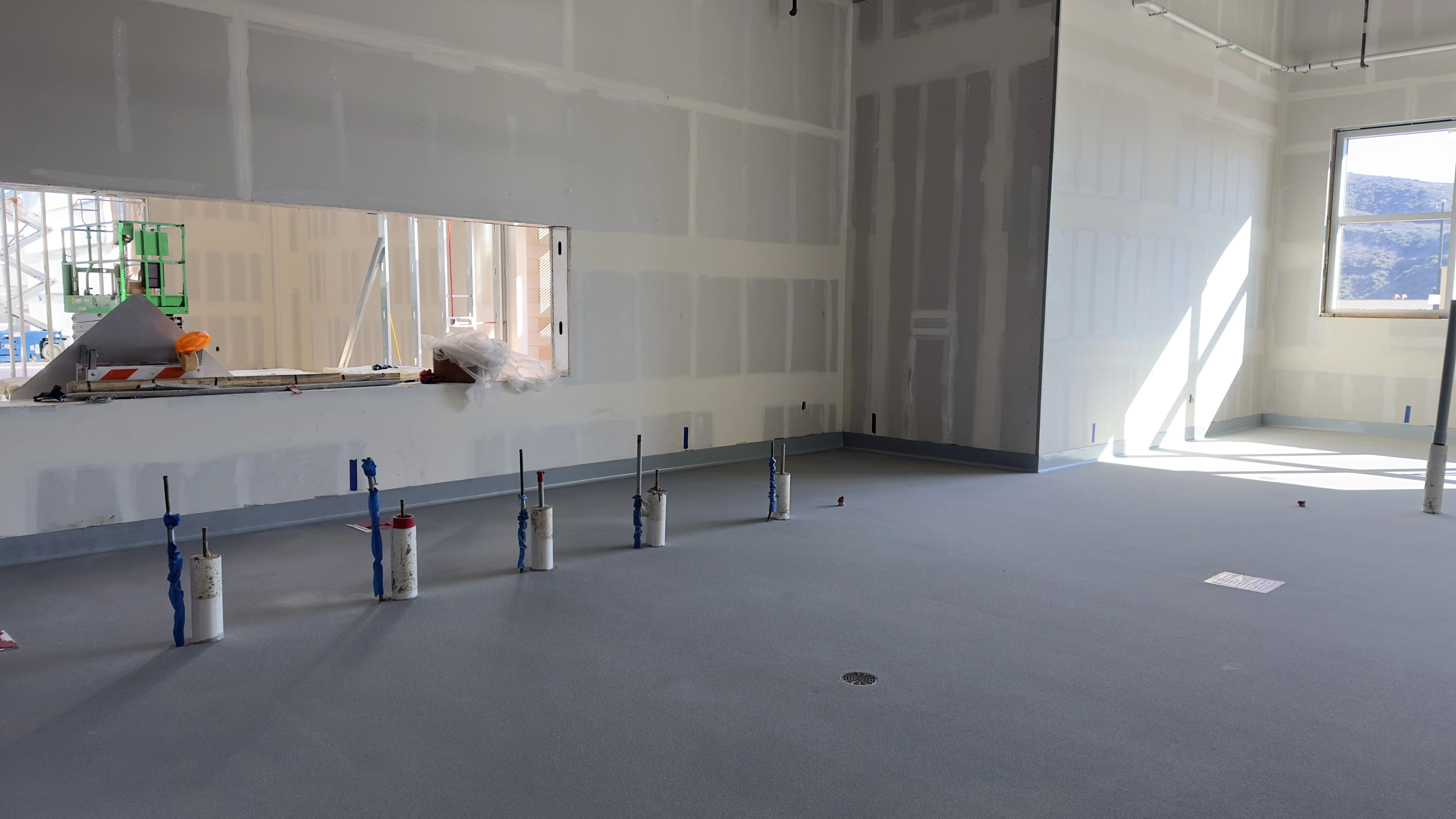 Unfinished interior room with exposed plumbing pipes, unpainted drywall, and a window letting in natural light; construction materials visible through a wall opening.