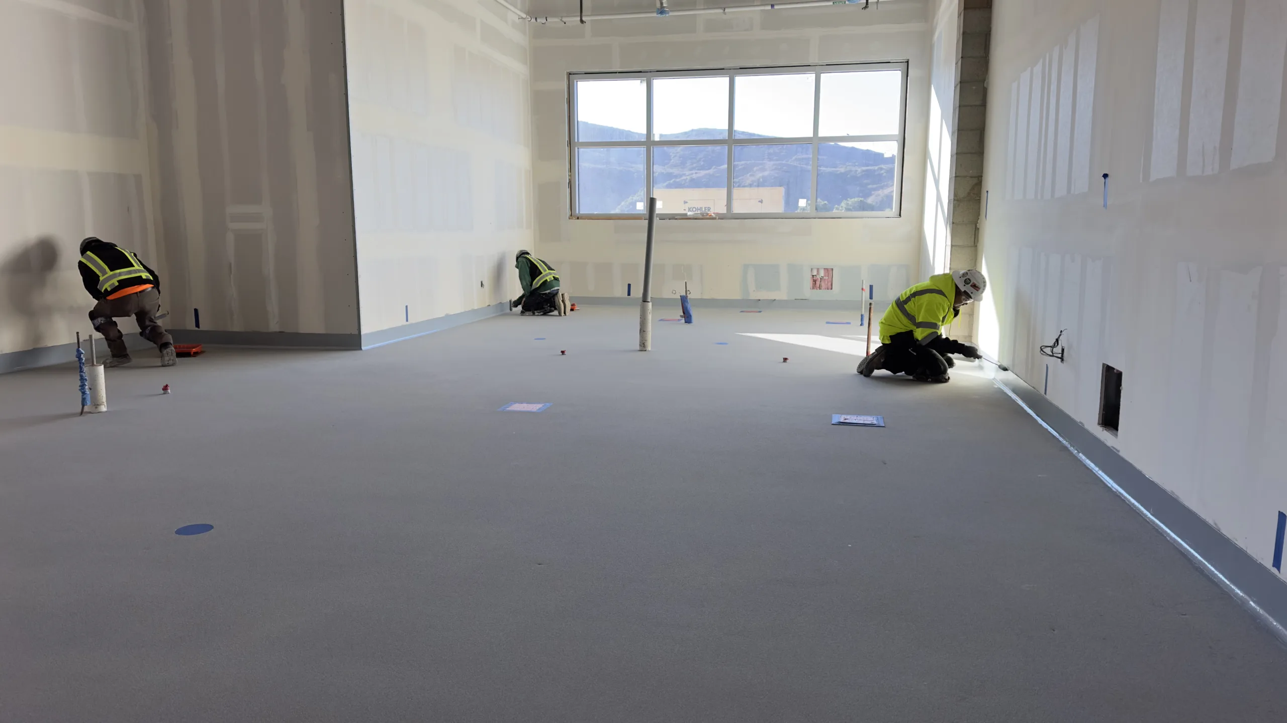 Three construction workers in safety gear work on the floor near the walls of a large, unfinished room with a window letting in sunlight.
