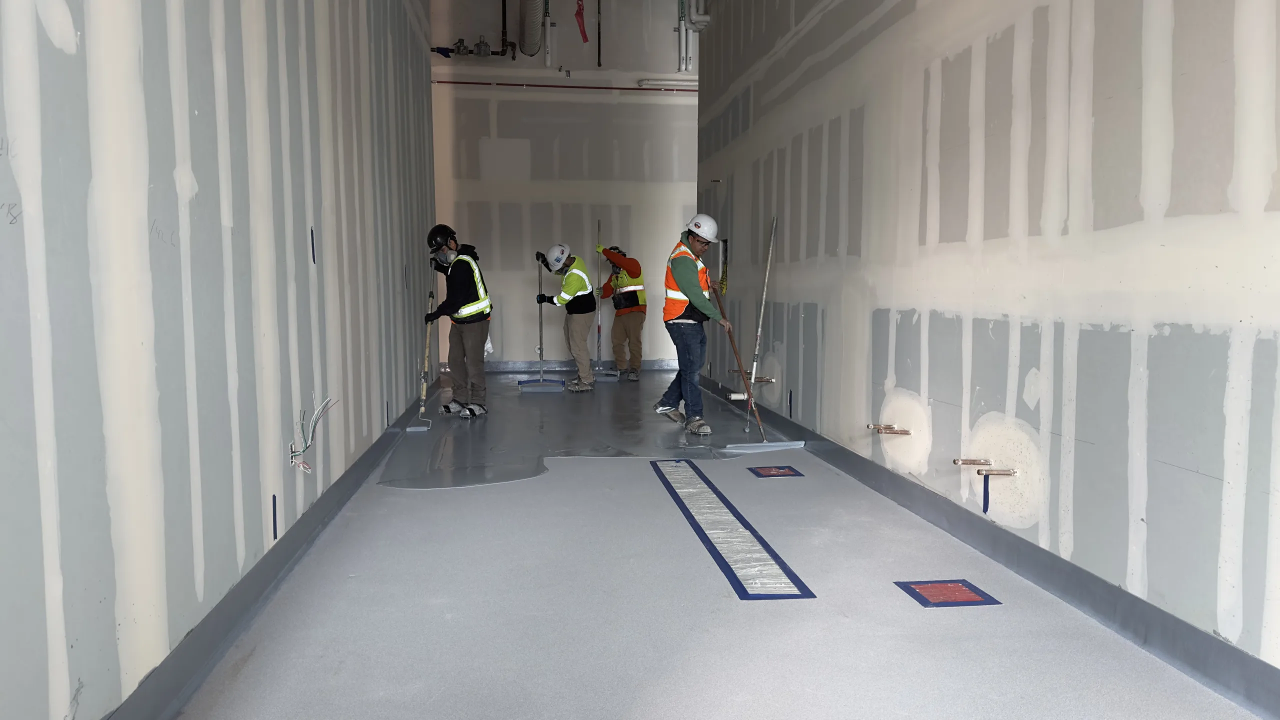 Four construction workers wearing safety gear apply coating to a large, unfinished room floor with taped-off sections and exposed pipes on the walls.