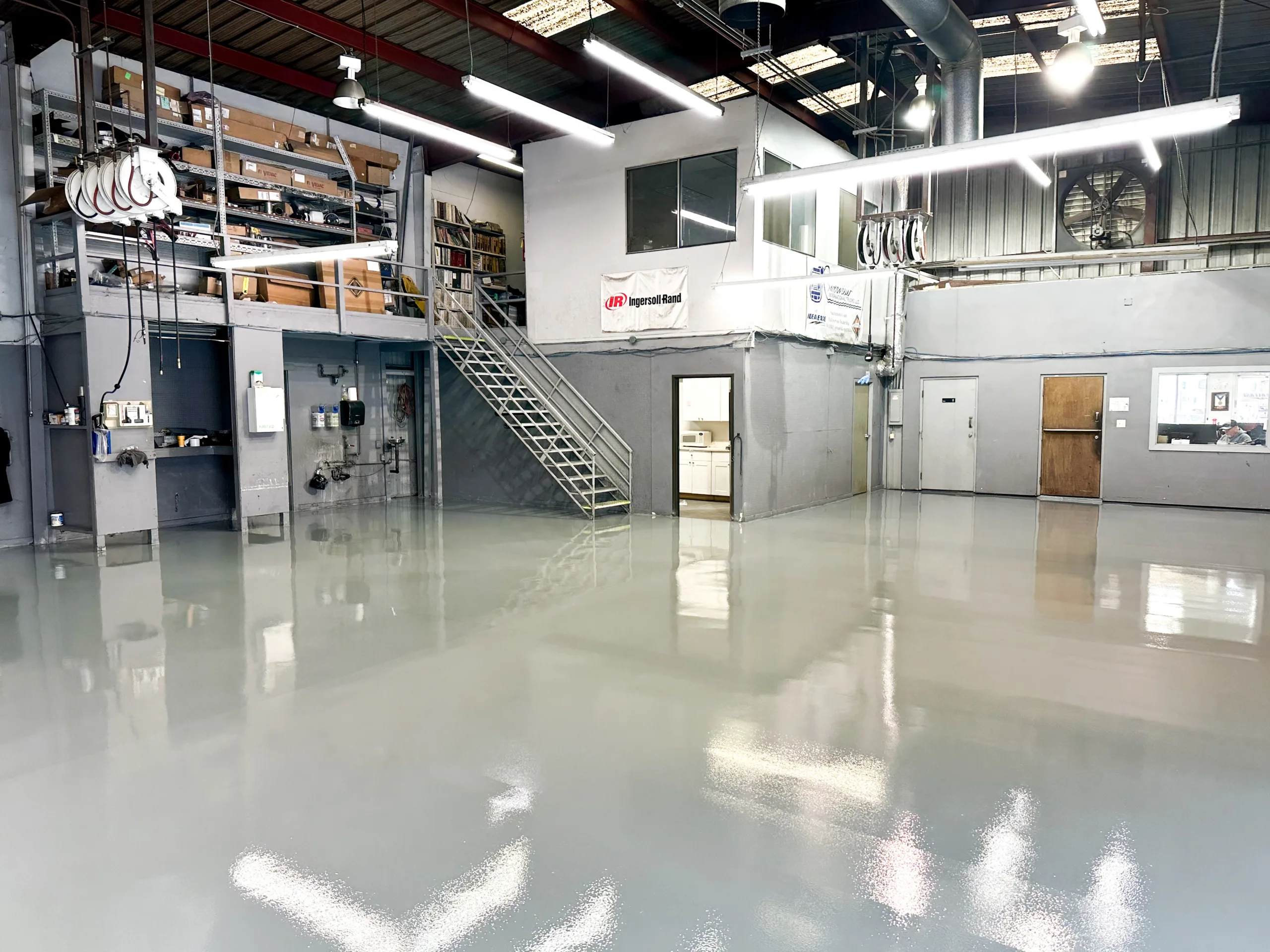 Spacious industrial warehouse with a polished concrete floor, metal staircase, shelves, and enclosed office spaces elevated above the main floor.