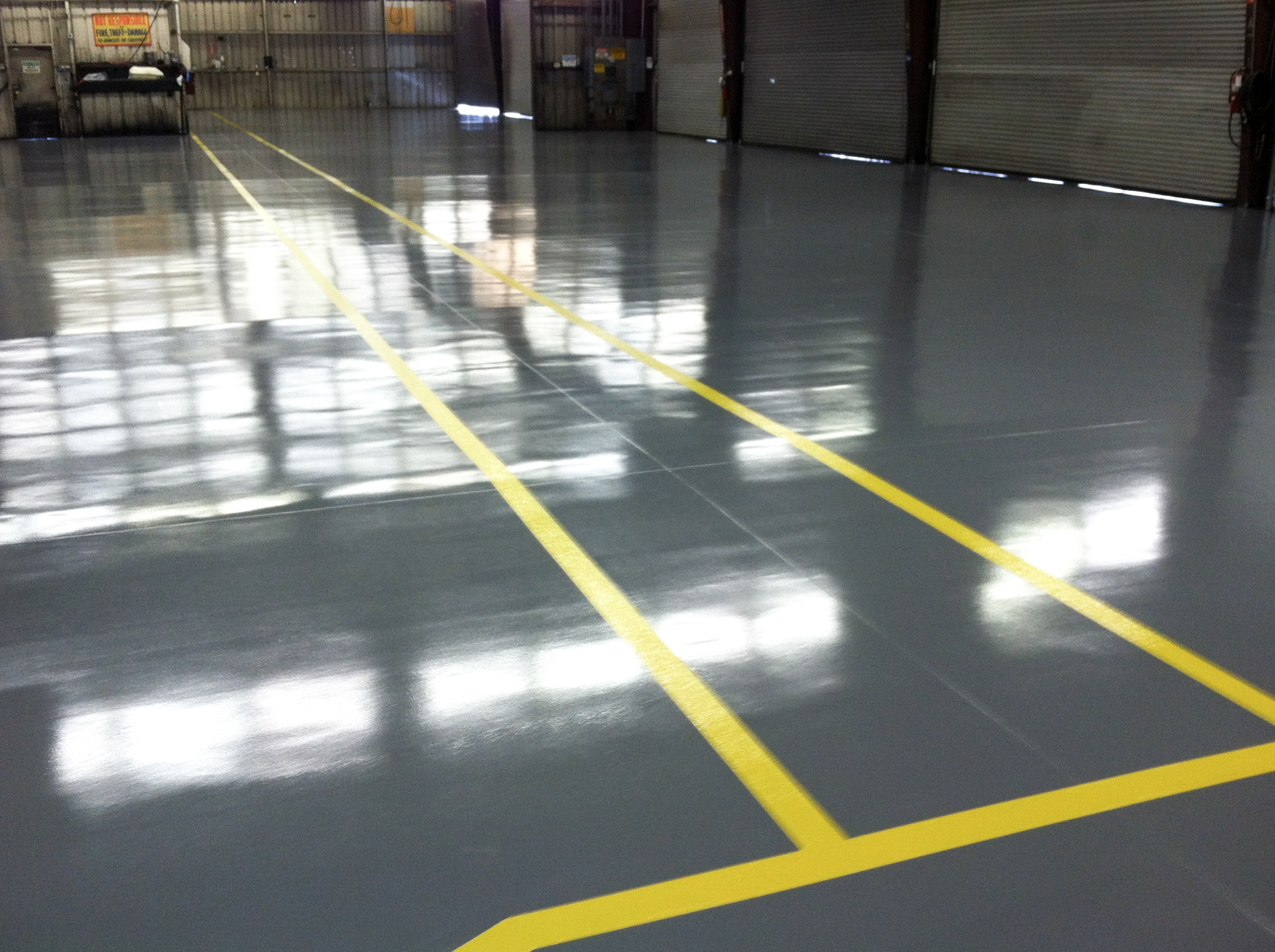 Large, empty warehouse with shiny gray epoxy flooring and bright yellow lines marking pathways. Metal walls and closed rolling doors are visible in the background.