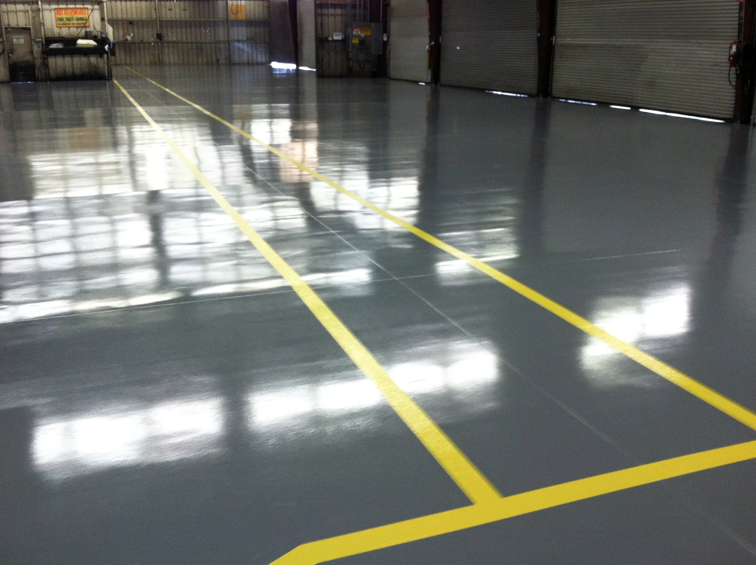 Large, empty warehouse with shiny gray epoxy flooring and bright yellow lines marking pathways. Metal walls and closed rolling doors are visible in the background.