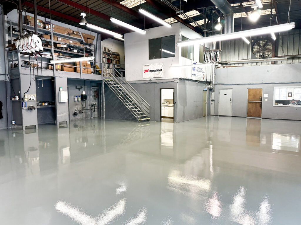 Large industrial workshop with gray polished floors, shelves with supplies, metal stairs to a loft area, and offices with windows and doors in the background.