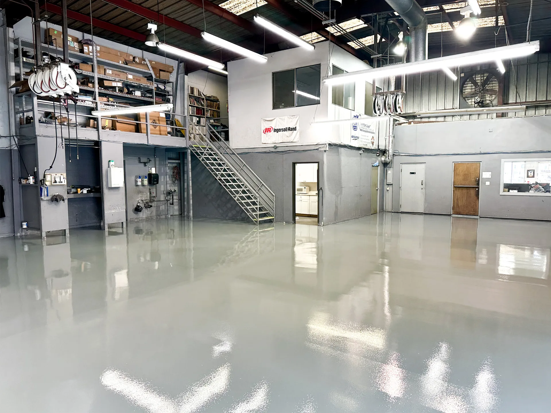 Large industrial workshop with gray polished floors, shelves with supplies, metal stairs to a loft area, and offices with windows and doors in the background.