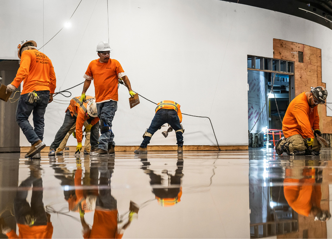 Five construction workers in orange shirts and safety gear are working on a shiny, reflective airport flooring inside a large building under construction.