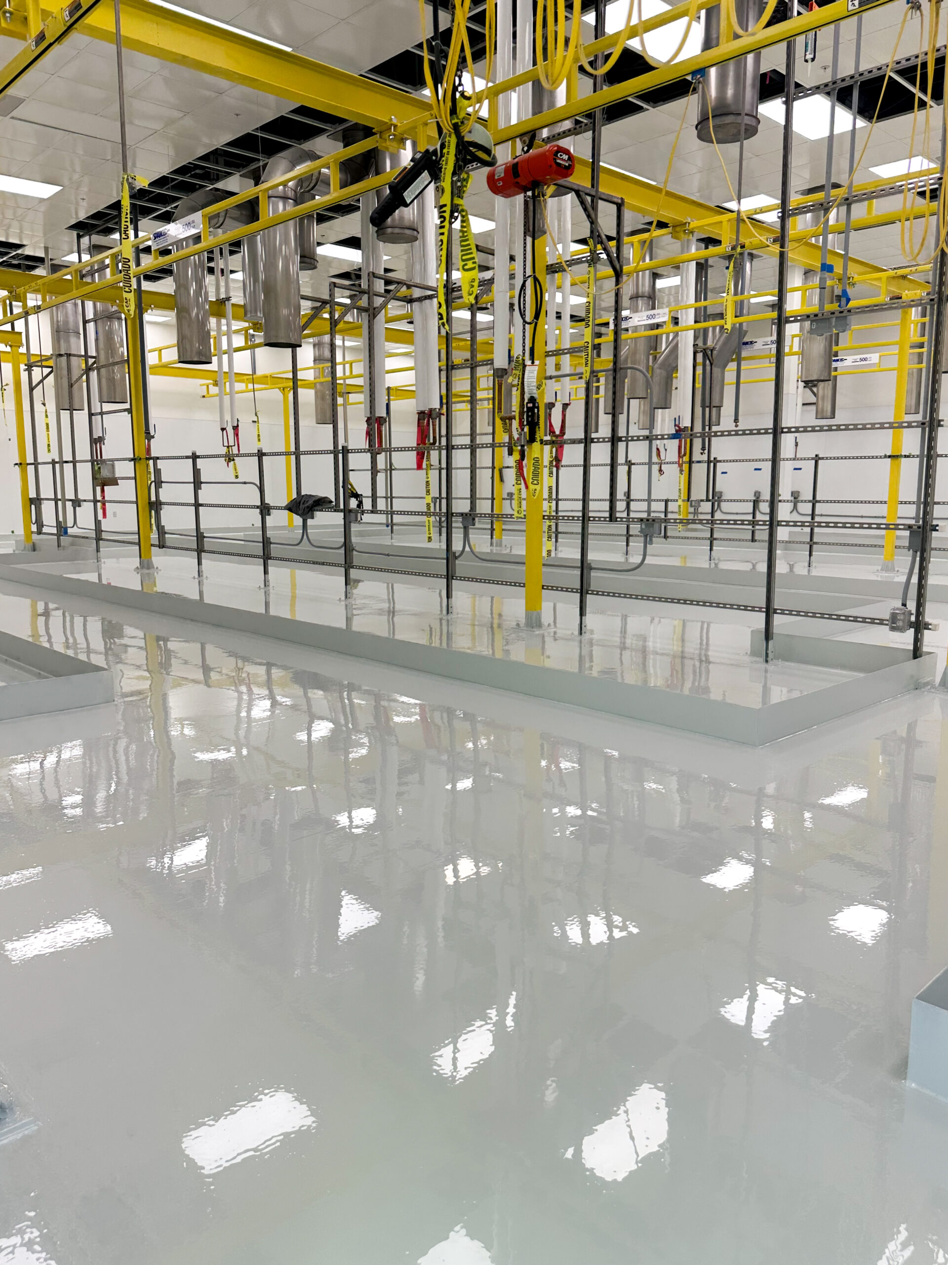 A clean industrial facility with a shiny white epoxy floor, yellow overhead pipes, suspended equipment, and metal railings.