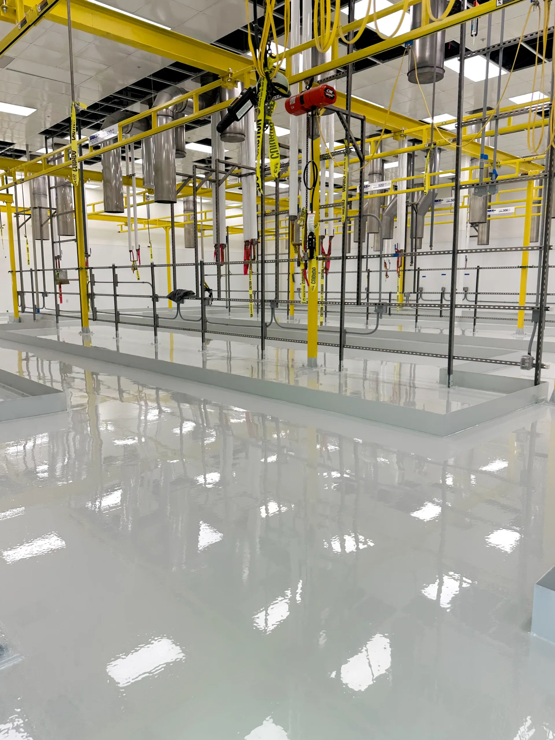 A clean industrial facility with a shiny white epoxy floor, yellow overhead pipes, suspended equipment, and metal railings.