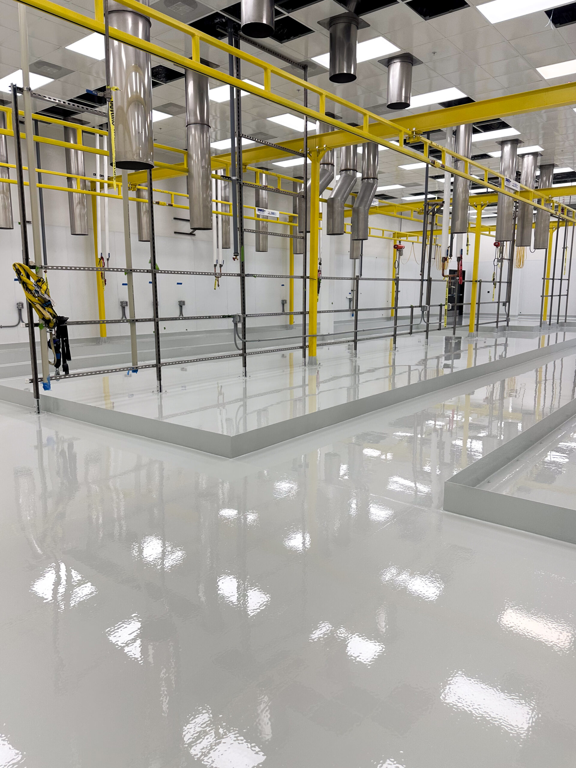 Clean, industrial facility with shiny white epoxy floors, yellow overhead rails, metallic vents, and bright ceiling lights reflecting on the surface.