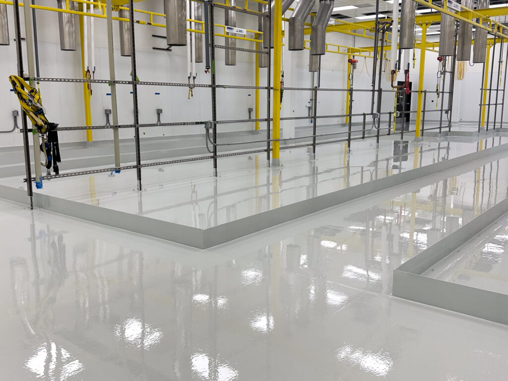 Industrial facility with freshly coated, glossy white epoxy floors, yellow support beams, metal pipes, and harness equipment along a wall.
