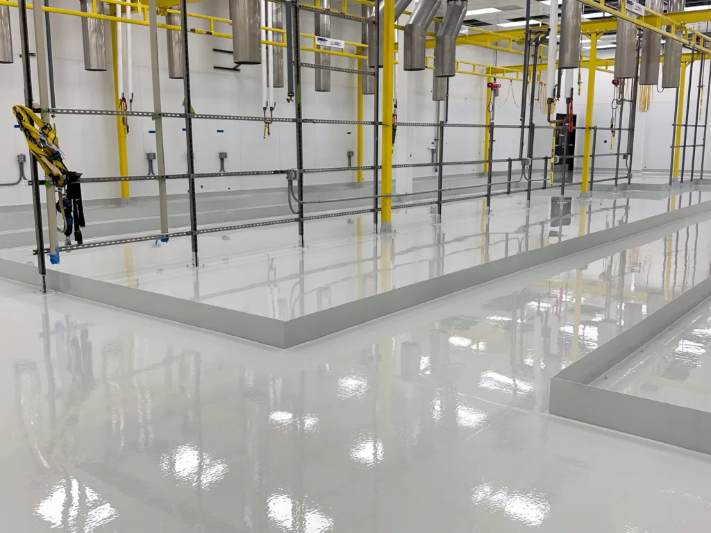 Industrial facility with freshly coated, glossy white epoxy floors, yellow support beams, metal pipes, and harness equipment along a wall.