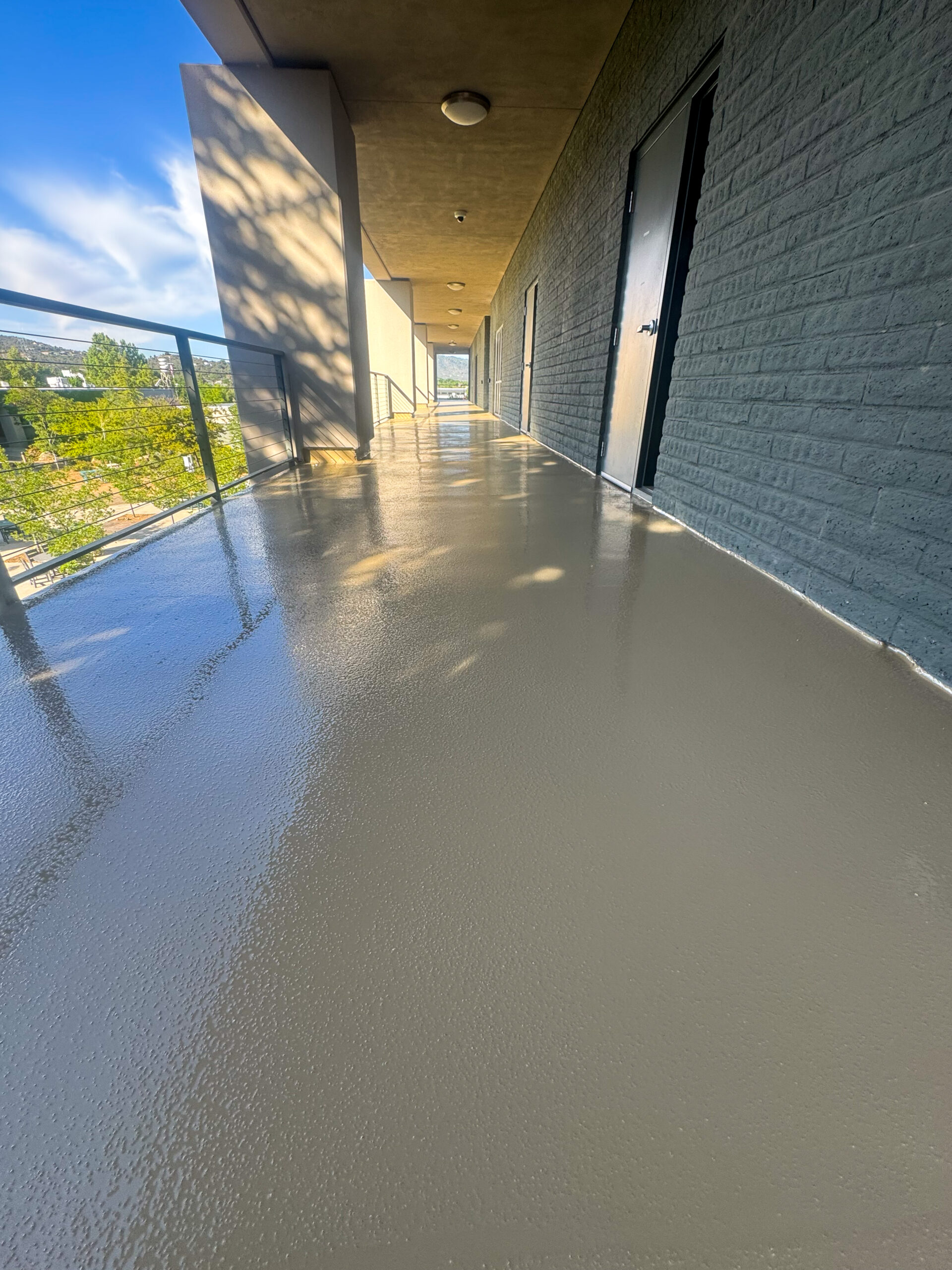 A freshly coated outdoor hallway with a smooth, shiny floor surface, bordered by a glass railing and gray brick wall with doors.