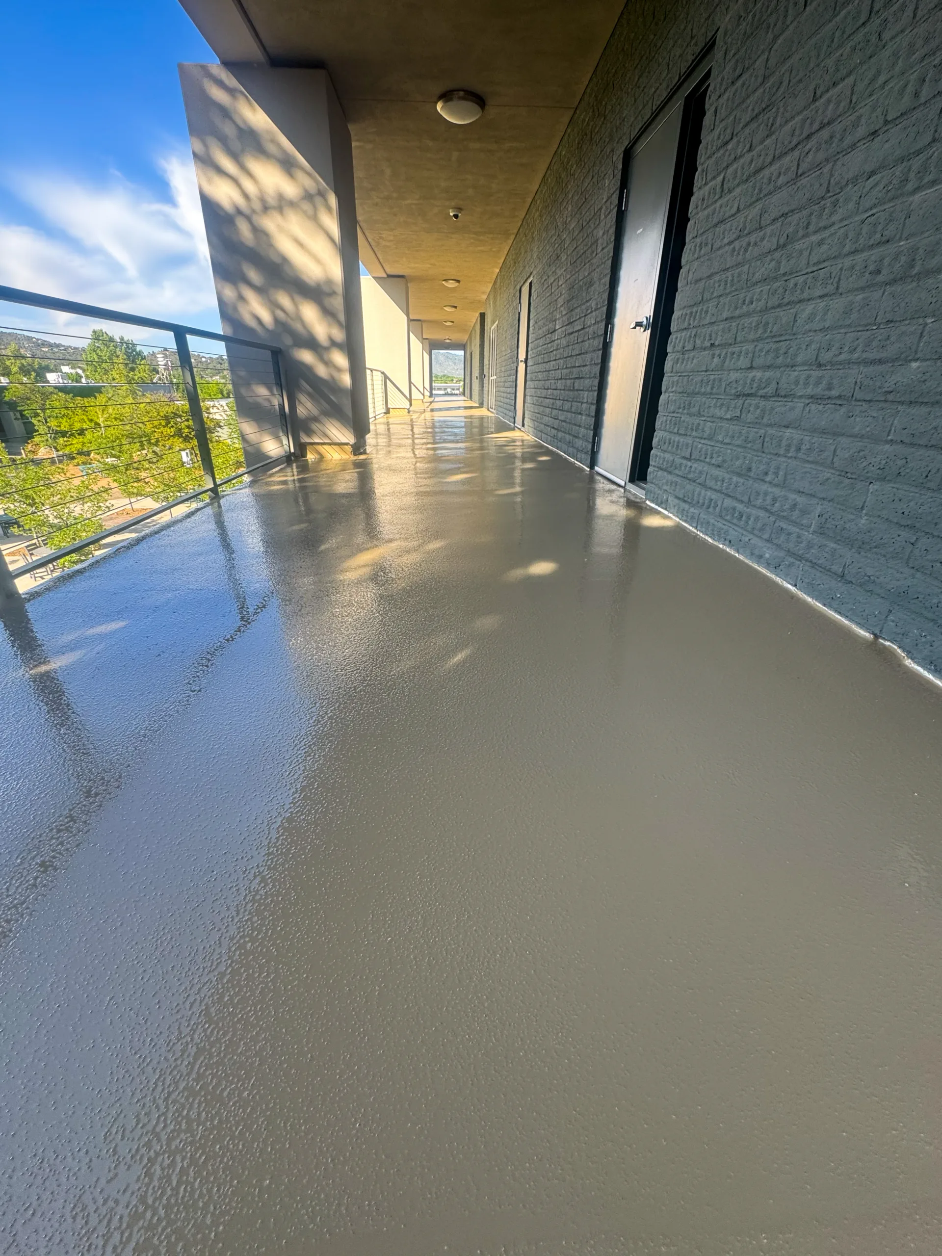 A freshly coated outdoor hallway with a smooth, shiny floor surface, bordered by a glass railing and gray brick wall with doors.