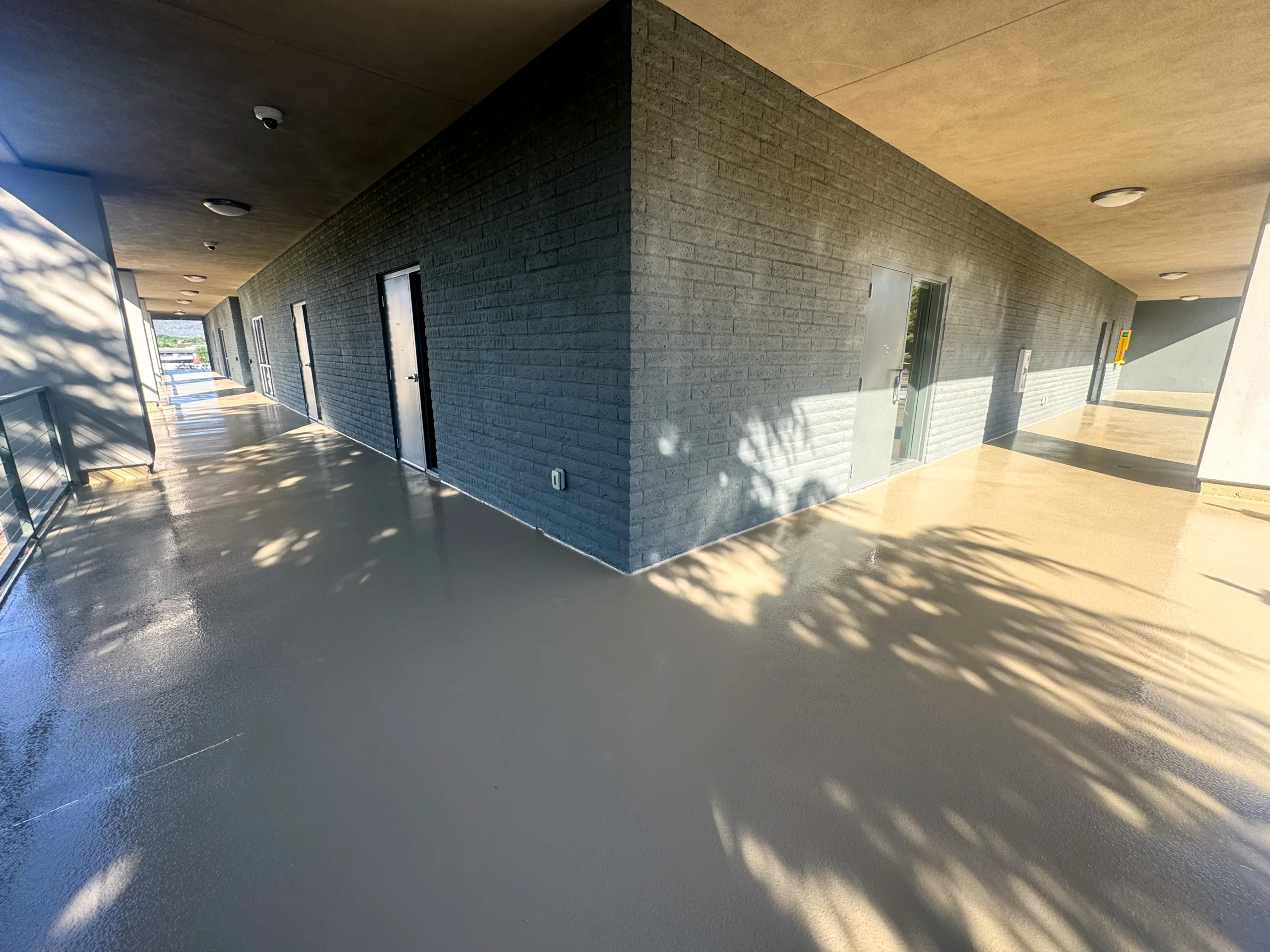 A clean, freshly-painted exterior corridor with several doors, modern lighting, and sunlight casting shadows on the floor and walls.
