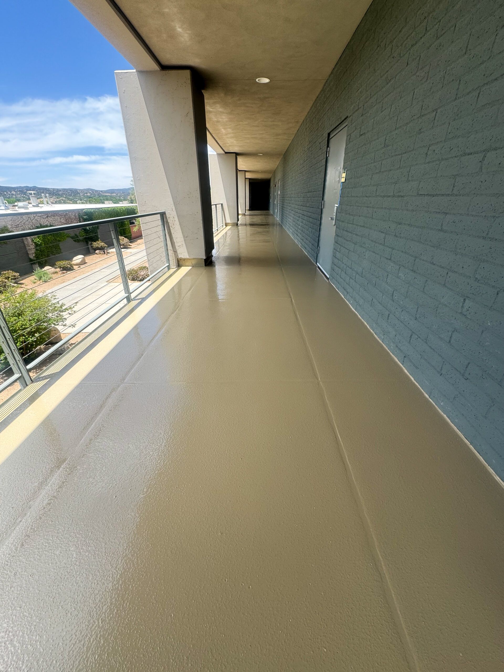 A long, clean outdoor hallway with a tan floor, gray brick wall on the right, and metal railing on the left, leading toward a dark doorway.
