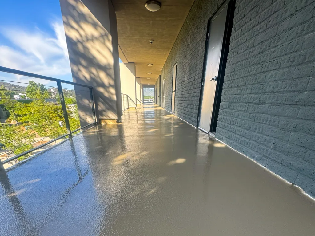 A clean, shiny outdoor walkway with a metal railing, concrete wall, and multiple black doors, photographed in daylight.