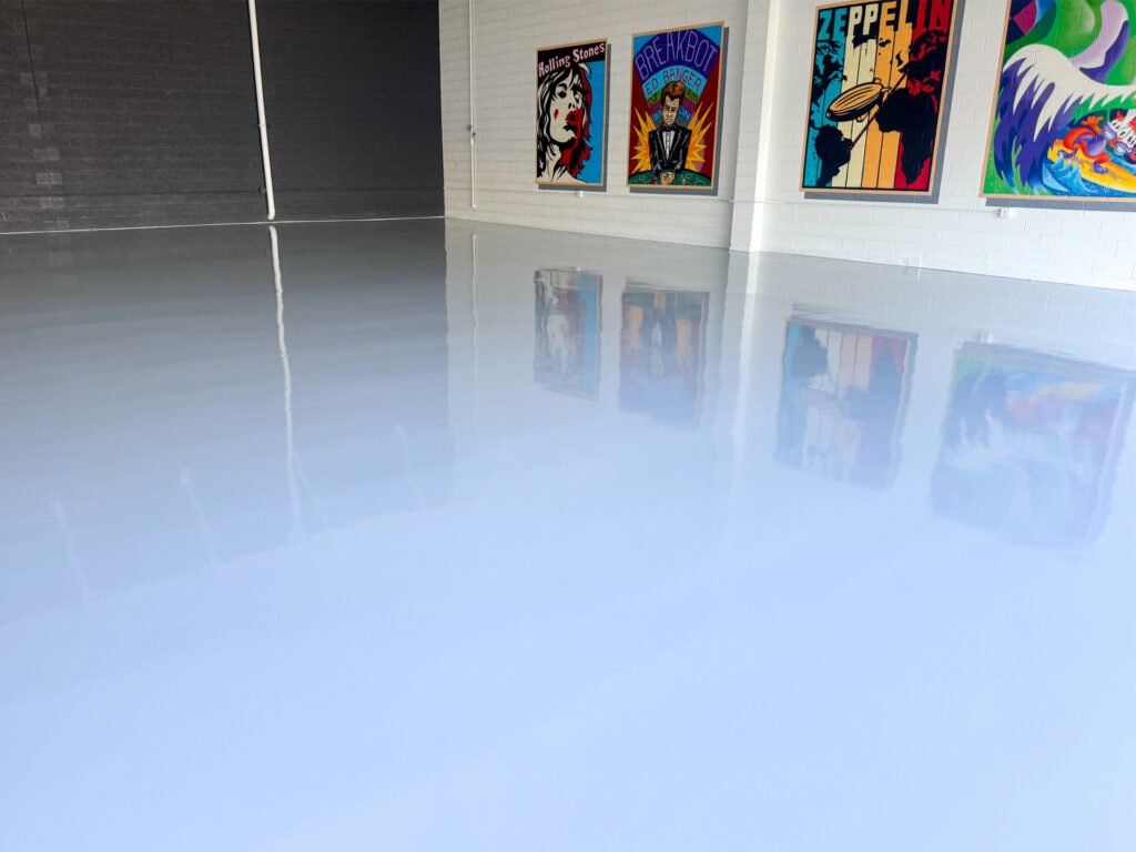 A smooth, glossy white floor reflects colorful framed posters on a white brick wall inside a spacious, modern room.