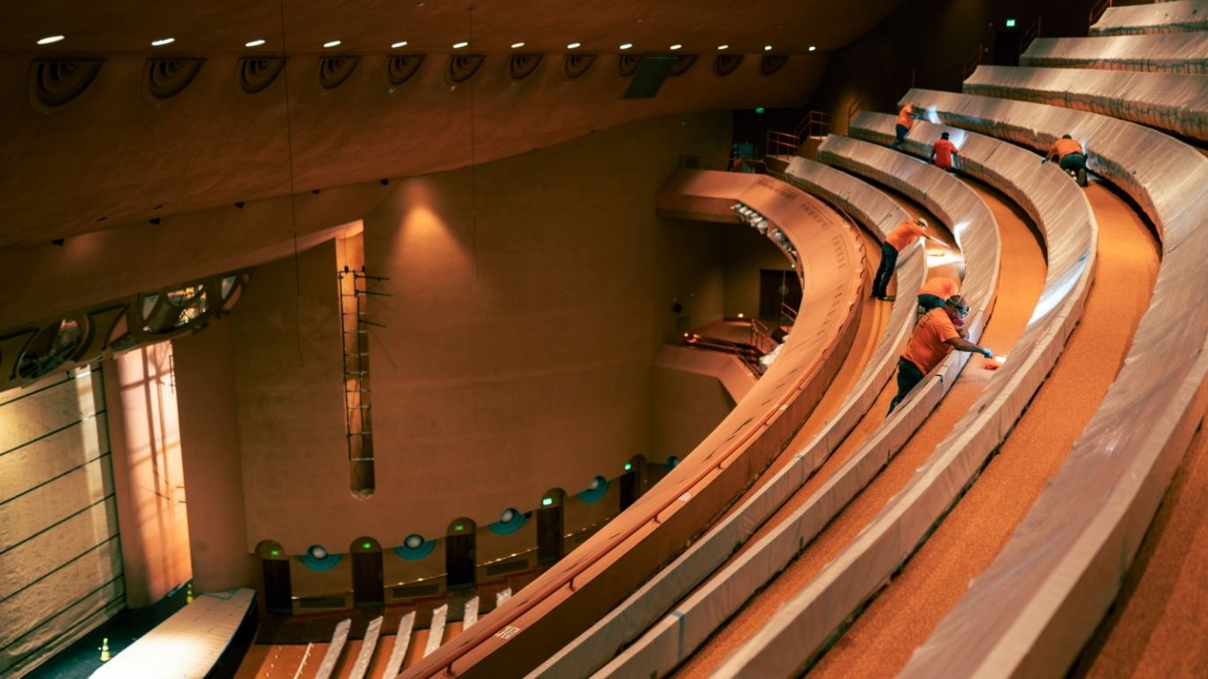 People are cleaning the curved, empty seats of a large indoor auditorium with warm lighting.
