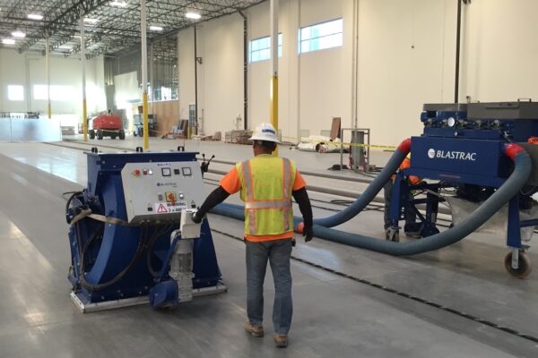 A construction worker wearing a safety vest operates a large floor grinding machine in a spacious, unfinished industrial building.