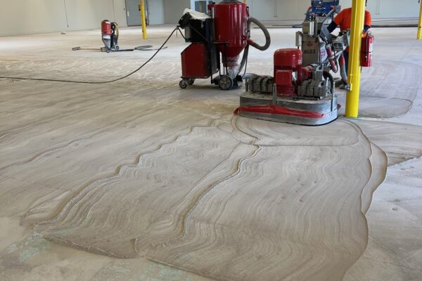 Workers operate industrial floor grinding machines to smooth a large concrete surface inside an open warehouse with exposed ceiling beams and yellow support columns.