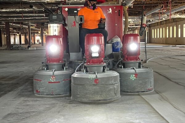 A construction worker operates a large ride-on concrete floor grinder with headlights on inside a spacious industrial building under construction.