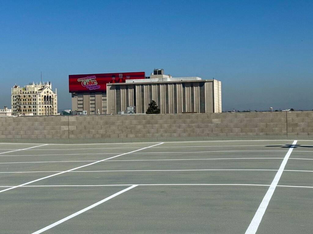 Empty rooftop parking lot with white parking lines, a low wall, and several buildings in the background, including a billboard advertising Coca-Cola. Clear blue sky overhead.