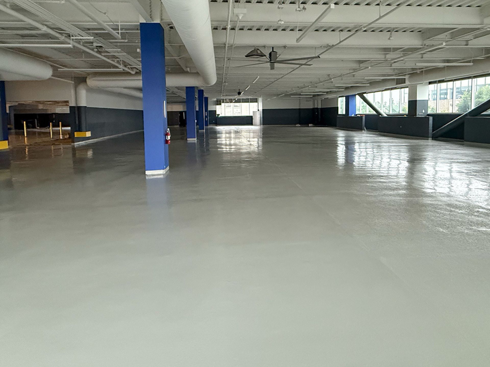 An empty indoor parking garage with smooth, freshly finished concrete floors, blue columns, windows on the right, and overhead lighting.