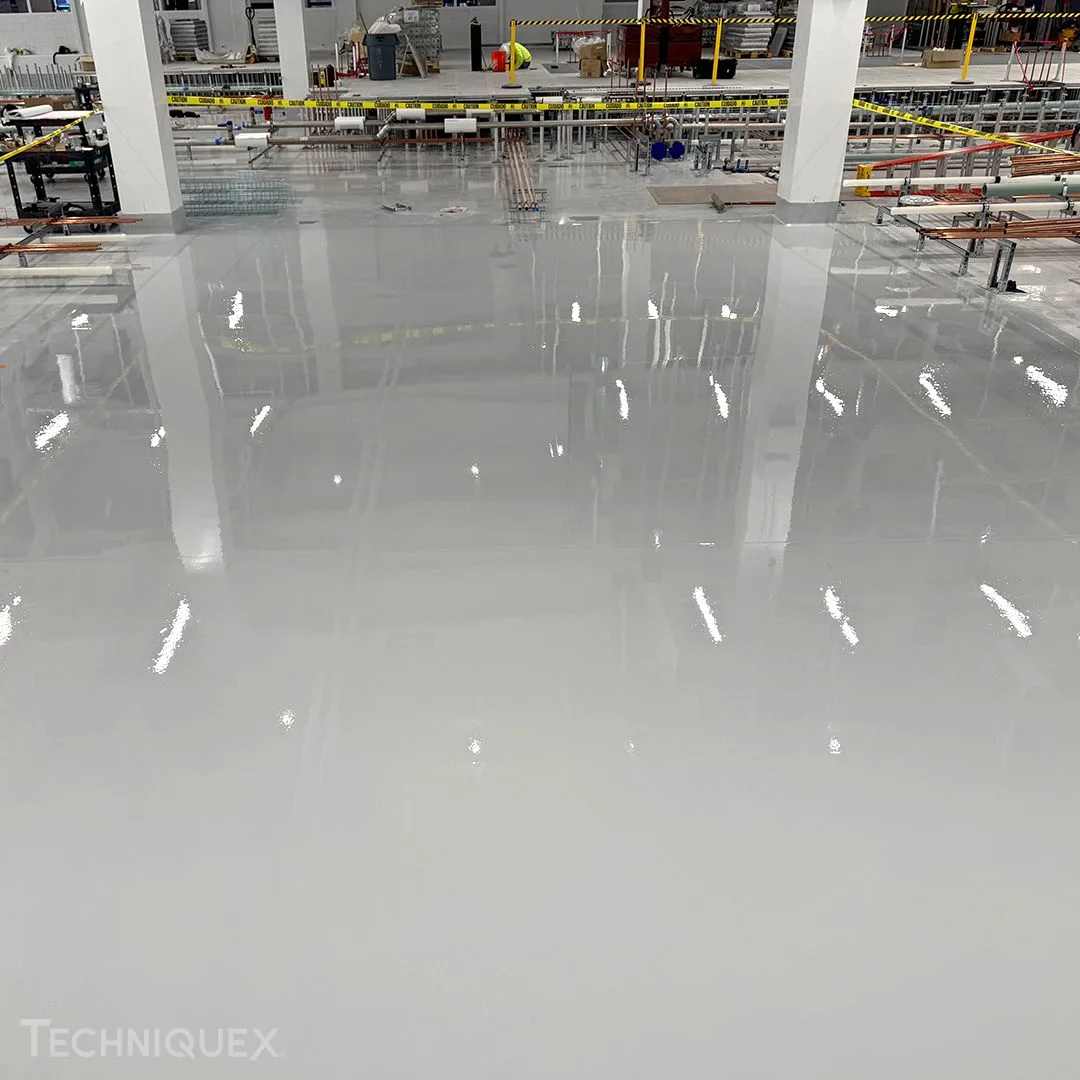 A large, freshly coated epoxy floor in an industrial facility displays a reflective surface and construction materials in the background, illustrating the differences between resin flooring vs epoxy flooring in such environments.
