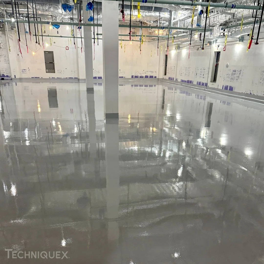 Large indoor room with a shiny, freshly coated epoxy floor—highlighting the appeal in the Resin Flooring vs Epoxy Flooring debate—white walls, exposed ceiling pipes, and various colored cords hanging down.